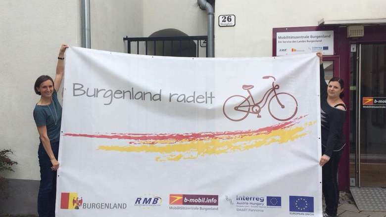 Leiterin der Mobilitätszentrale Burgenland Christine Zopf-Renner  zeigt mit der Mitarbeiterin Lisa Liszt den neuen Projektbanner